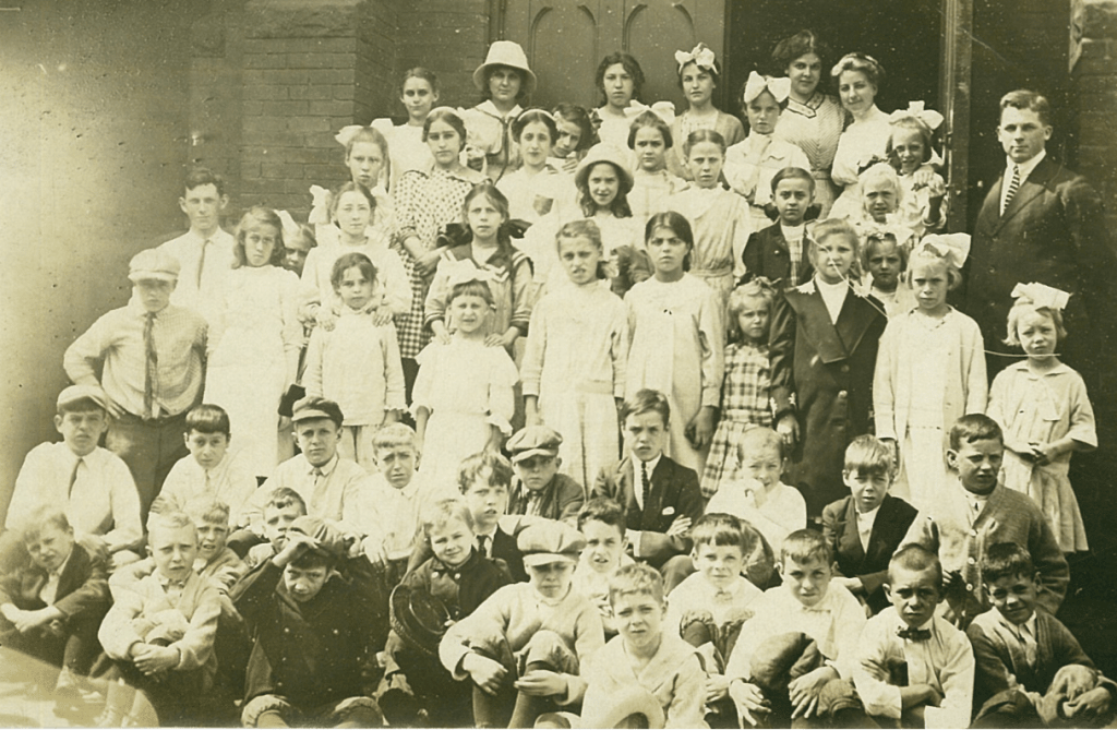 Several dozen children pose together on a staircase. An adult man in a suit stands to the right. A wooden door behind the group stands open.