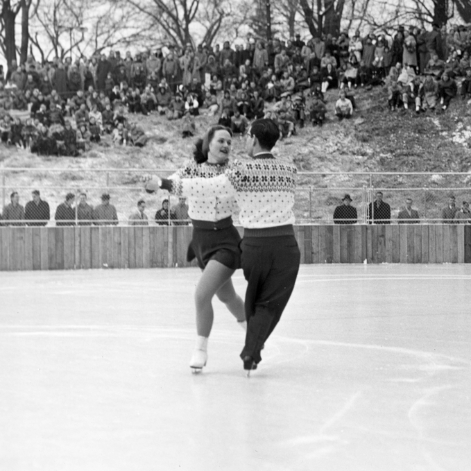 A crowd sits on a grassy hill, and watches two figure skaters perform a dance on the ice rink. The pair wear matching sweaters, and are holding hands with their left arms extended.