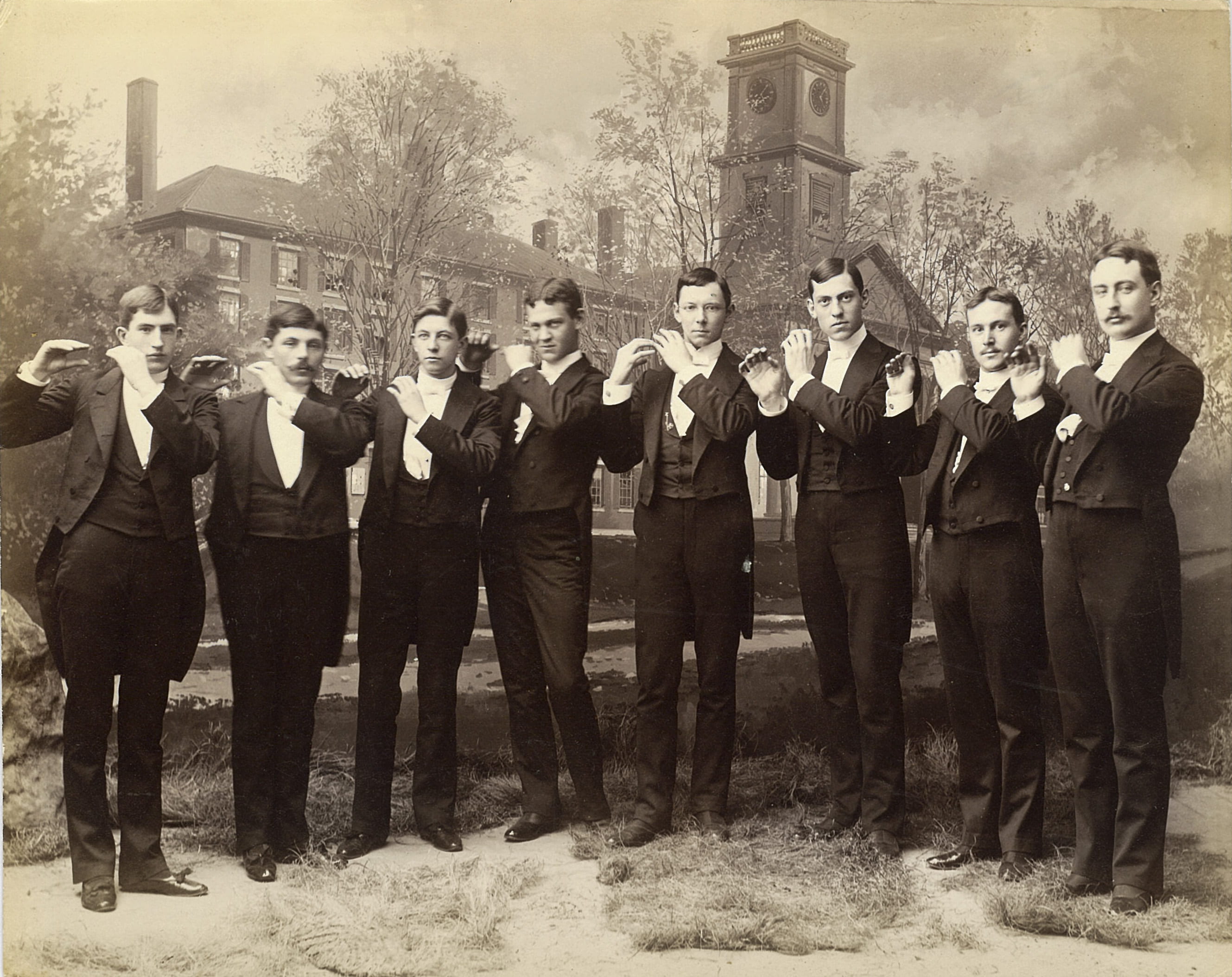 A photograph of eight students in formal wear in front of a painted backdrop of Johnson Chapel. The students appear to be holding invisible flutes.