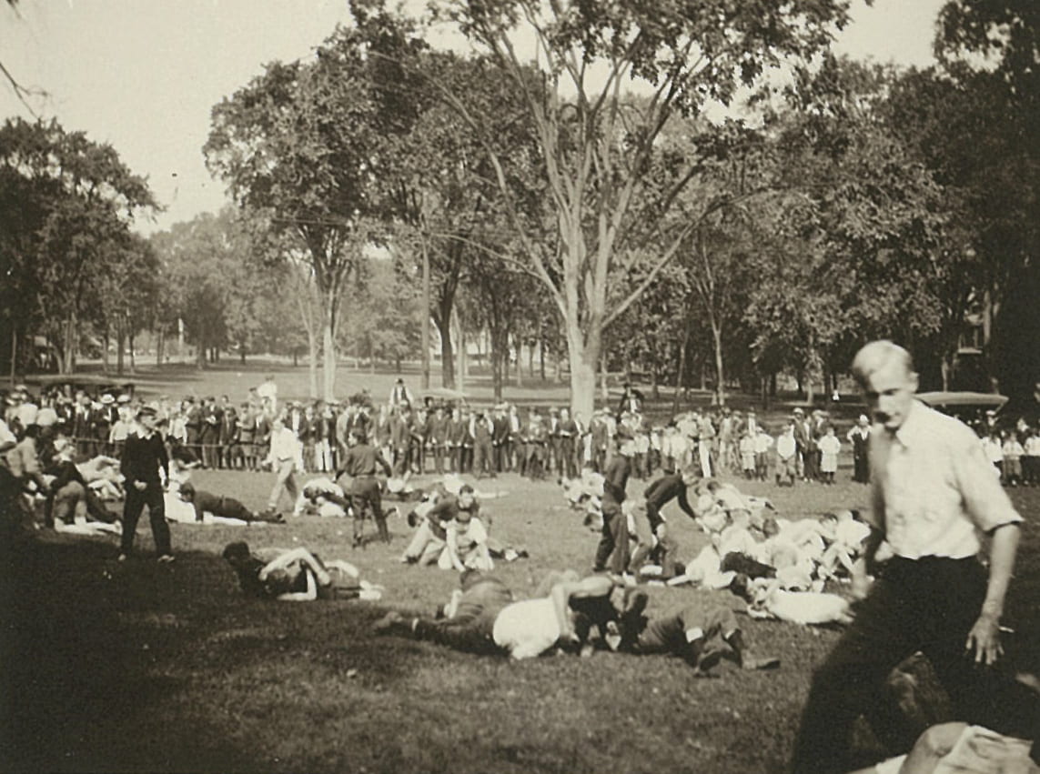 Photograph of clusters of students rolling on the ground in fisticuffs, behind them is a crown of onlookers behind a rope.