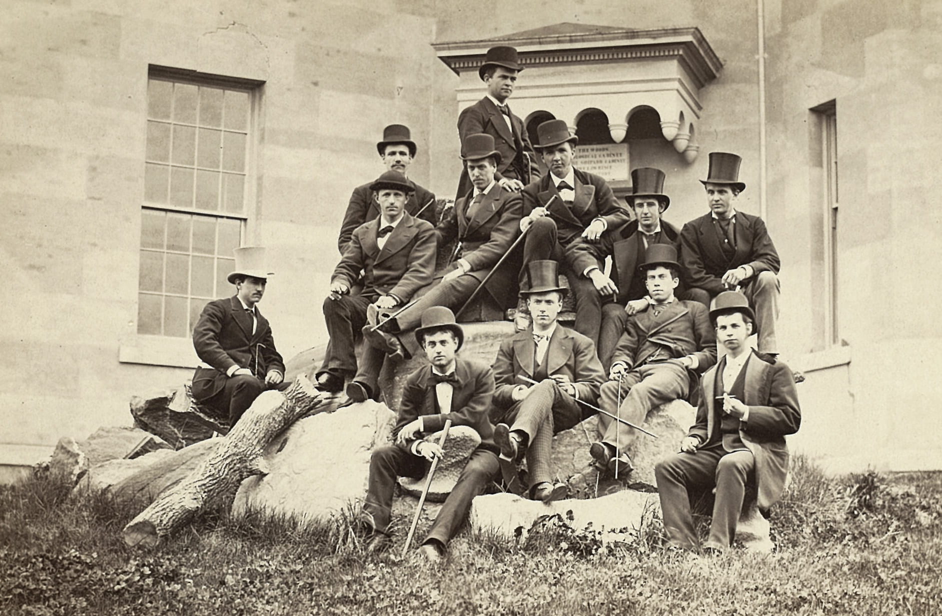 Photograph of a group of students with canes and top hats sitting on a large rock, probably from the 1880s.