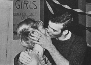 A black and white photograph of a man and a woman kissing at a party. Both are dressed according to traditional gender norms.